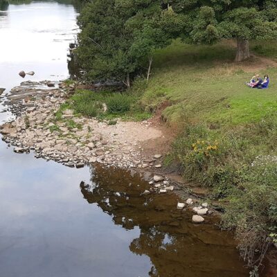 An aerial view of a wide a slow moving section of the river Eden. A stone a boulder shelf encroaches into the river on the inner bank of a slight bend. To the right a small grass bank with two people having a picnic.