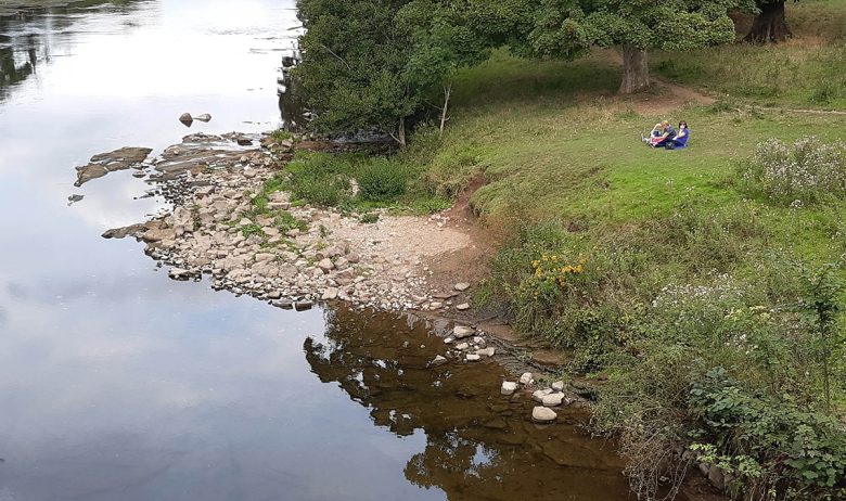 An aerial view of a wide a slow moving section of the river Eden. A stone a boulder shelf encroaches into the river on the inner bank of a slight bend. To the right a small grass bank with two people having a picnic.