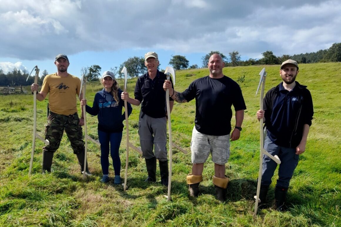 Five people stood in open grass field each holding a large wooden scythe.
