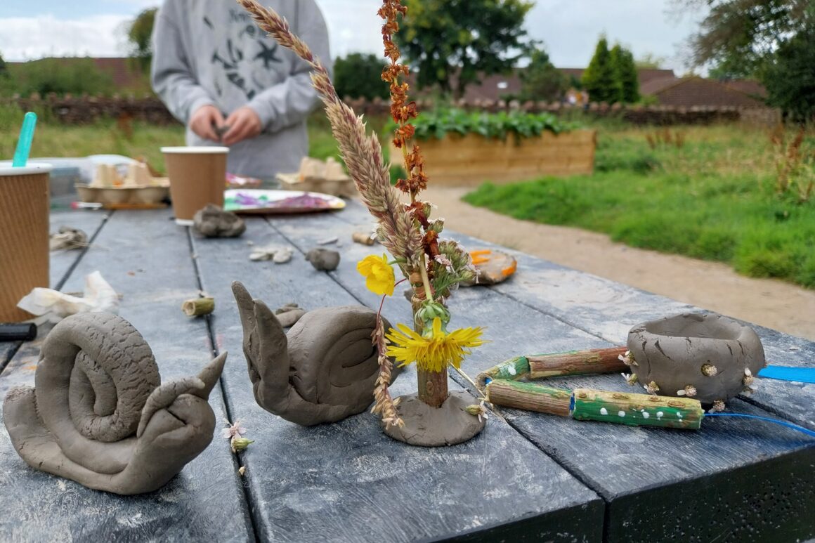 Plasticine snails and flowers in a vase sit on a wooden picnic table on a gravel path