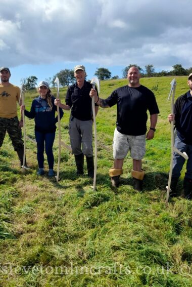 Five people stood in open grass field each holding a large wooden scythe.