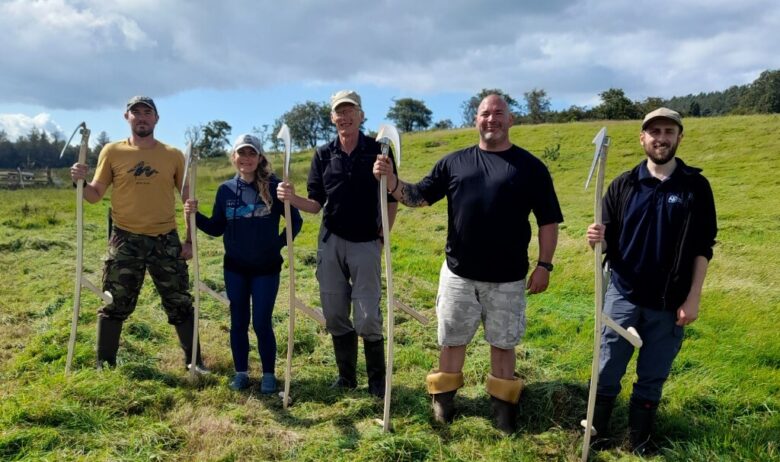 Five people stood in open grass field each holding a large wooden scythe.