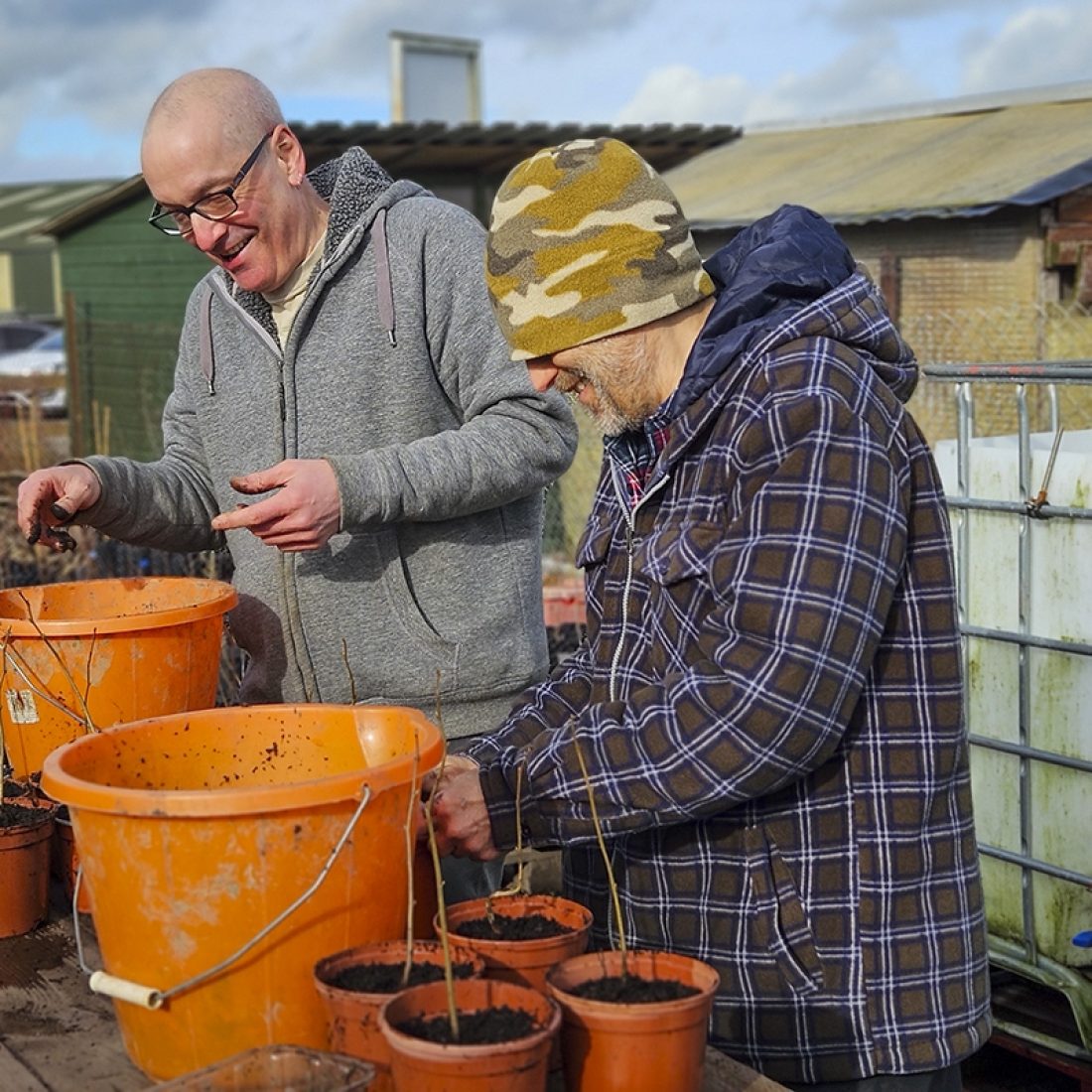 Two volunteers at a wooden table, putting small saplings into individual pots, filled with soil decanted from large orange buckets sat on the table.