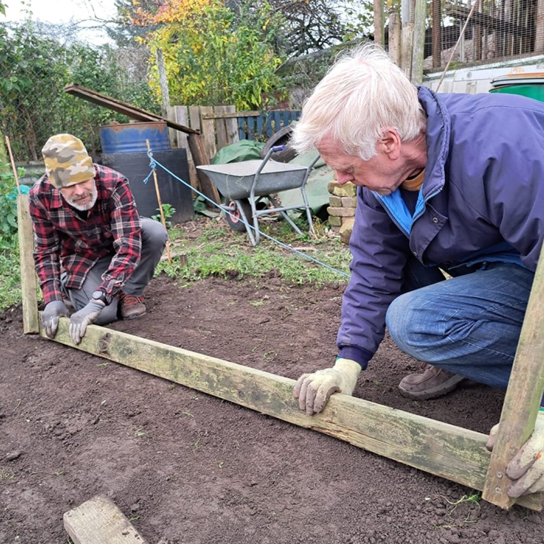 Two volunteers running a long straight wooden plank across loose bare soil to level it off in readiness for the erection of a polytunnel on the site.