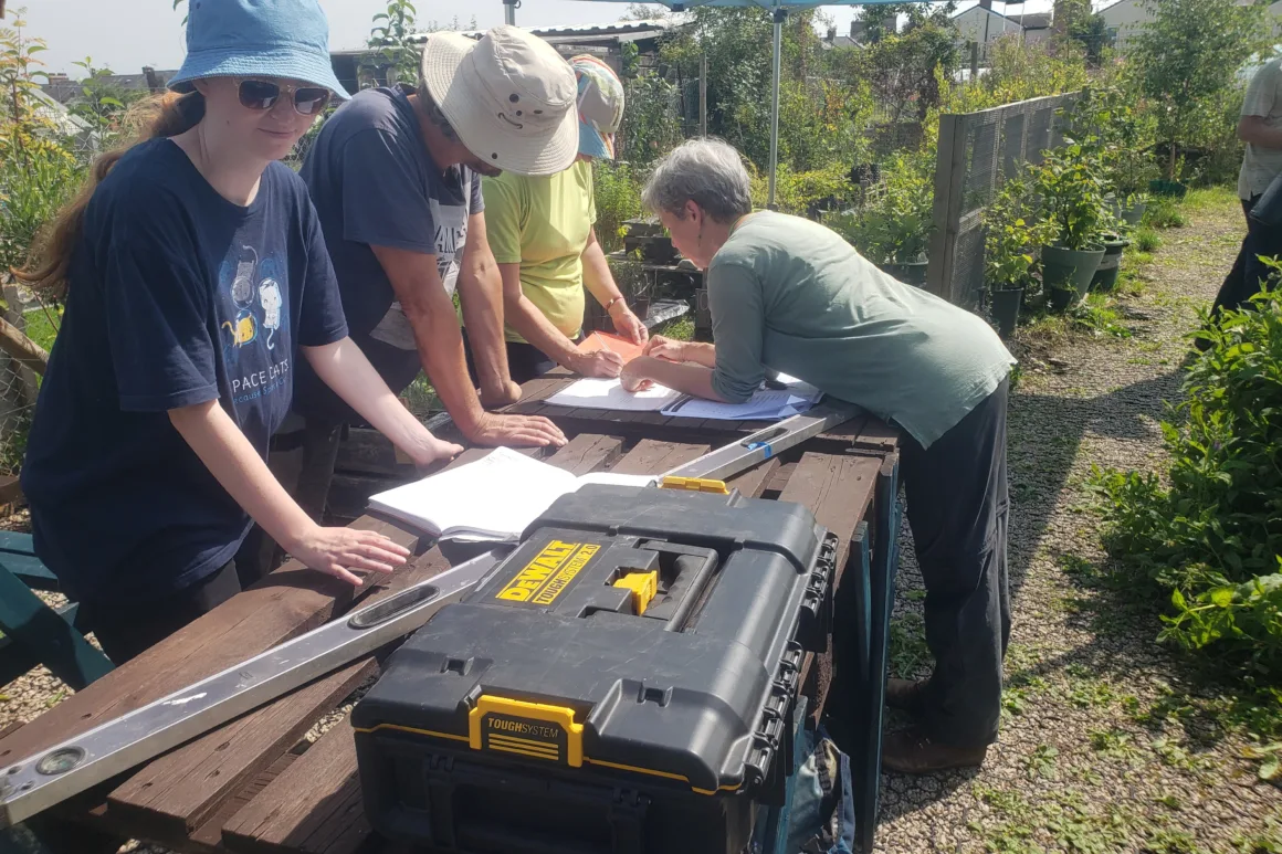 Four volunteers gathered around an outdoor 'potting' table in sunshine looking at building plans with spirit level and toolbox in foreground. Lots of young saplings in separate pots in the background.