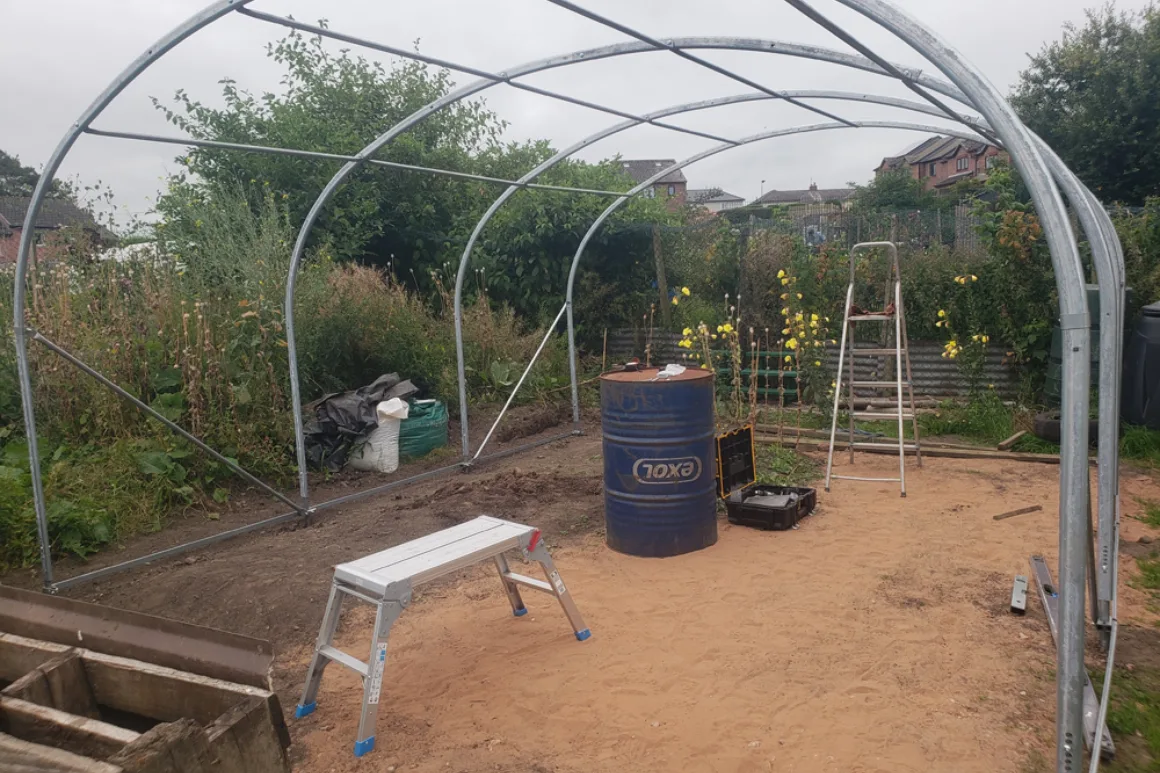 A flat section of sandy ground with the tubular metal frame of a large polytunnel structure spanning the space. Outdoor setting with small saplings in pots surrounding the bare ground where the frame sits.