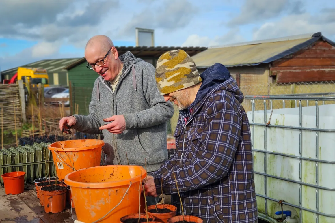 Two volunteers at a wooden table, putting small saplings into individual pots, filled with soil decanted from large orange buckets sat on the table.