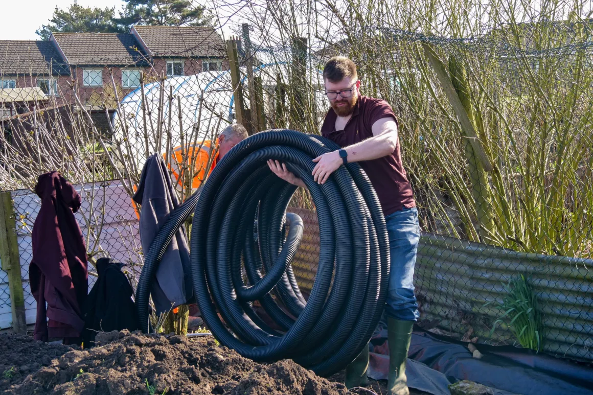 A volunteer moving a large coil of plastic water pipe into position, ready to be laid out in a trench dug around the border of the Penrith community tree nursery.
