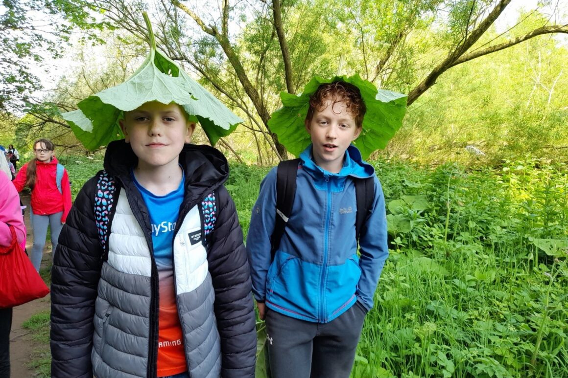 A group of young people in a woodland setting posing for a photograph. Two young people in the foreground have fashioned hats from large leaves found in the woodland.