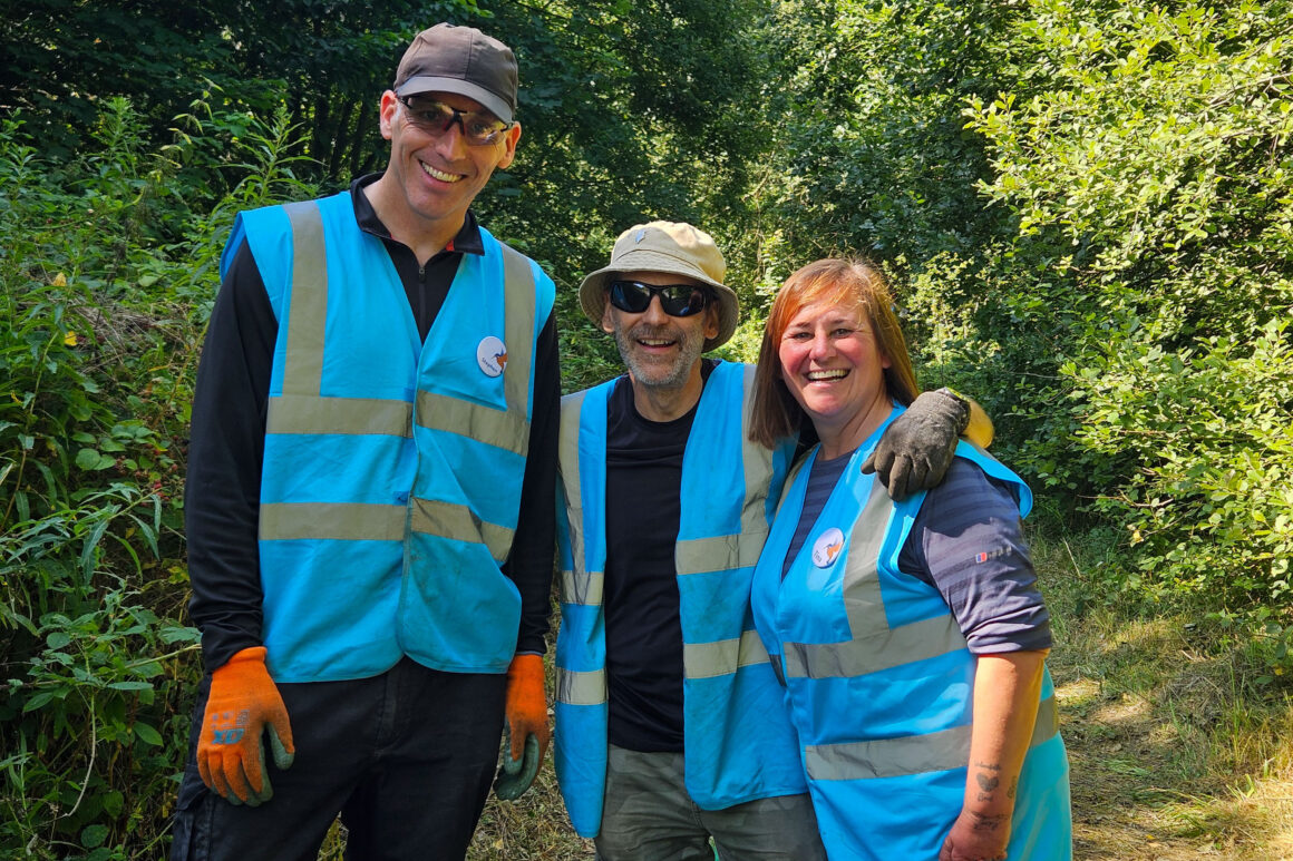 Three people in blue high visibility jackets and gardening gloves and sunglasses posing for an image on a grassy pathway in front of trees in summer leaf.