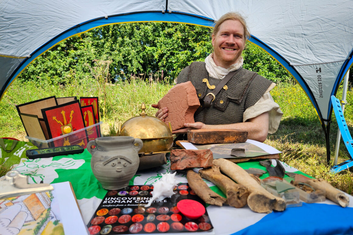 A man dressed in roman costume sat behind a table covered in roman artefacts including pottery and helmets. Roman shields in red with gold decoration sit behind. All under an awning on green grass.