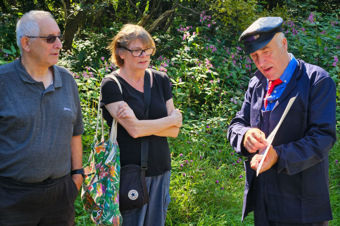 Two people being shown information on a board by a man dressed in British Rail uniform from the 1960's; consisting of dark blue overalls, dark blue jacket, red tie and blue railway cap.