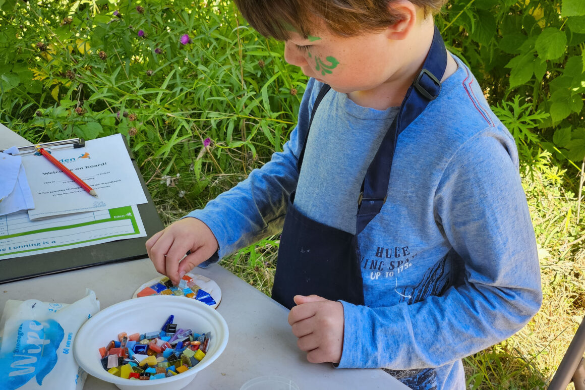 A young person at a table choosing coloured pieces of mosaic from a bowl to arrange on a colourful tile, creating artwork.