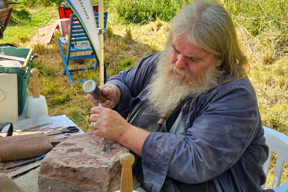 A bearded man in blue overalls sat at a table. Using a chisel and hammer to carve a design into a large piece of sandstone.