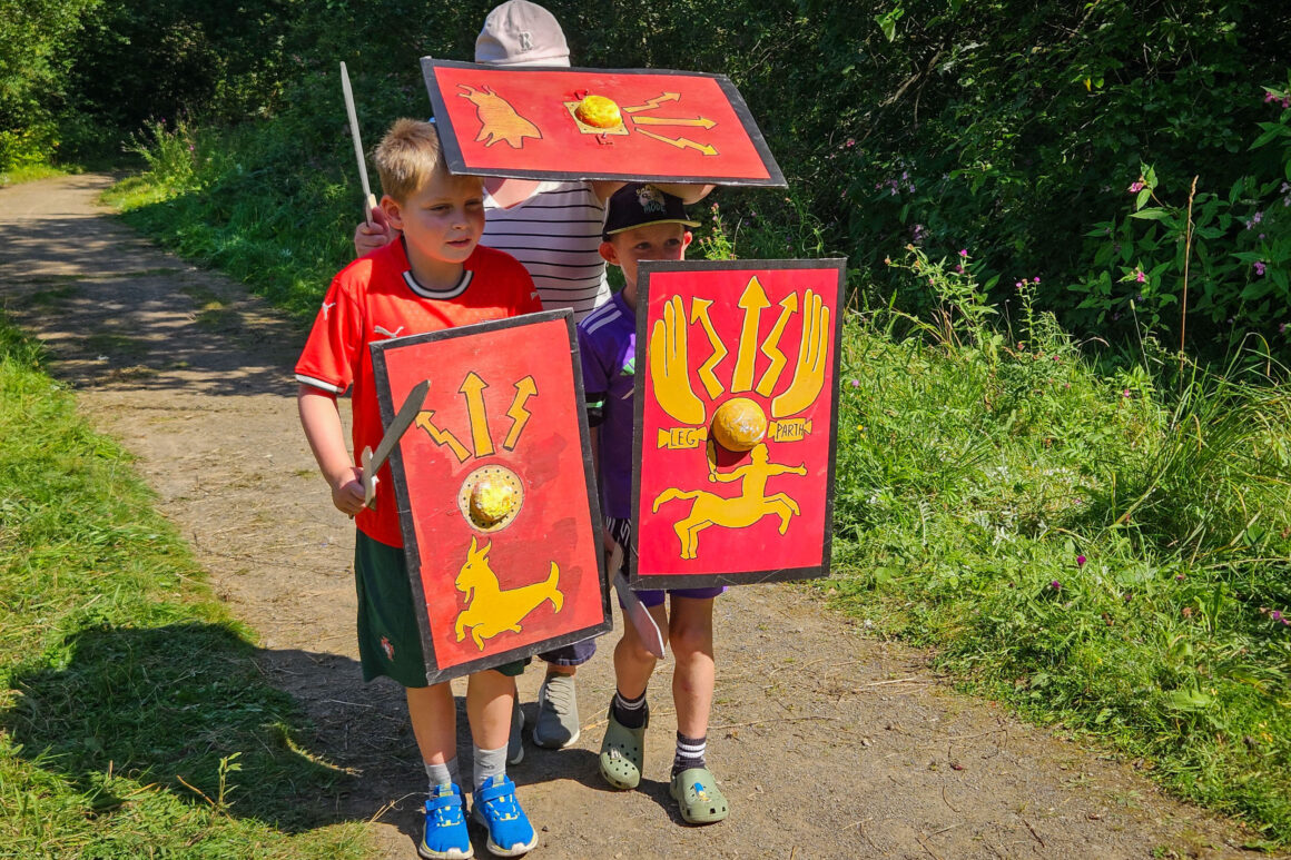 Three young people stood together on a gravel path holding replicas of roman shields and carrying toy swords.