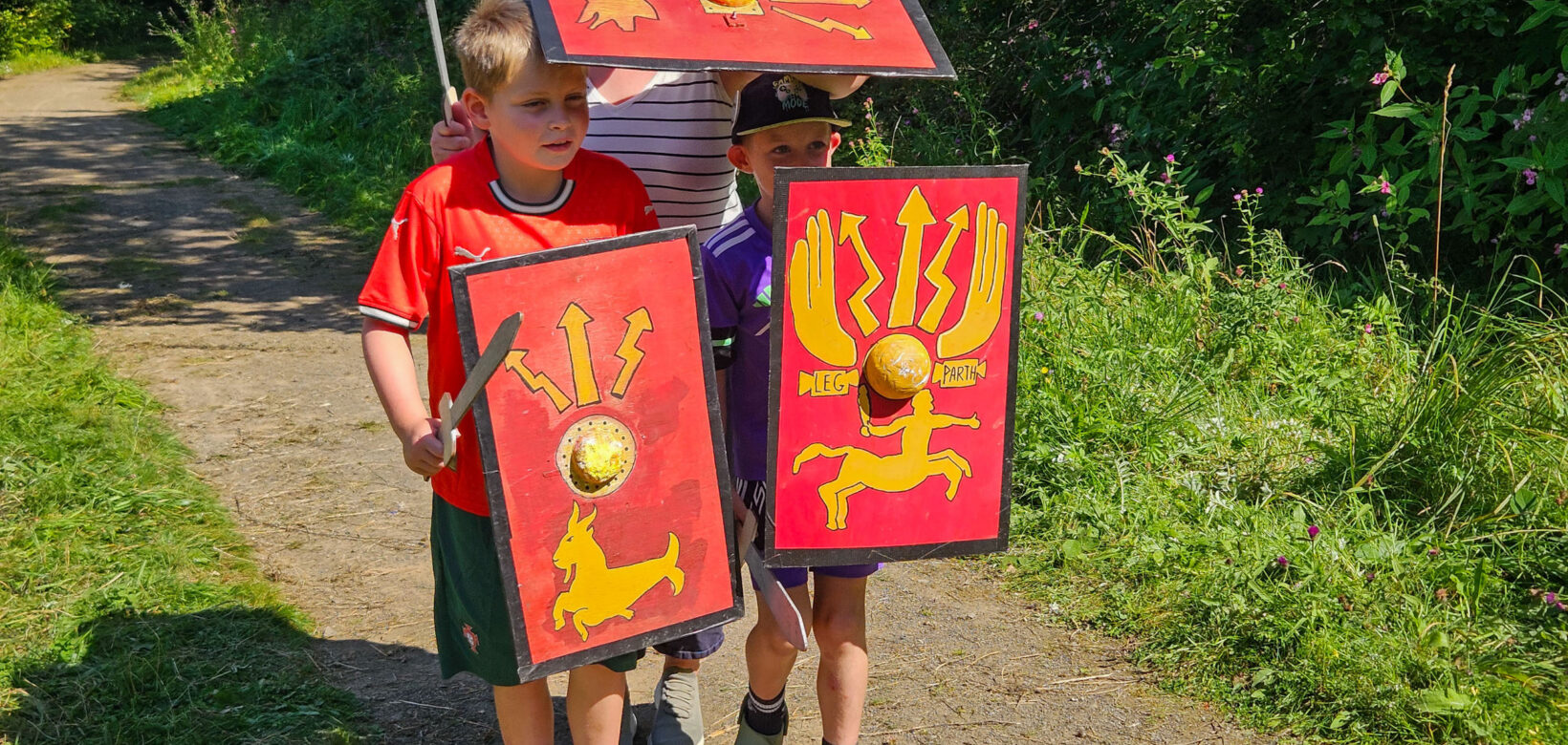 Three young people stood together on a gravel path holding replicas of roman shields and carrying toy swords.