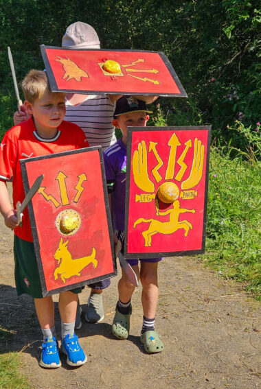 Three young people stood together on a gravel path holding replicas of roman shields and carrying toy swords.