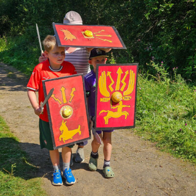 Three young people stood together on a gravel path holding replicas of roman shields and carrying toy swords.
