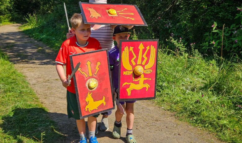 Three young people stood together on a gravel path holding replicas of roman shields and carrying toy swords.
