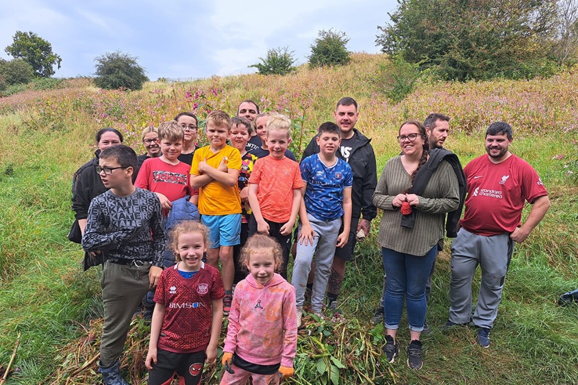 A group of young people in colourful T shirts are stood on a large pile of Himalayan balsam collected as part of a sponsored balsam bash held at Petteril Vale.