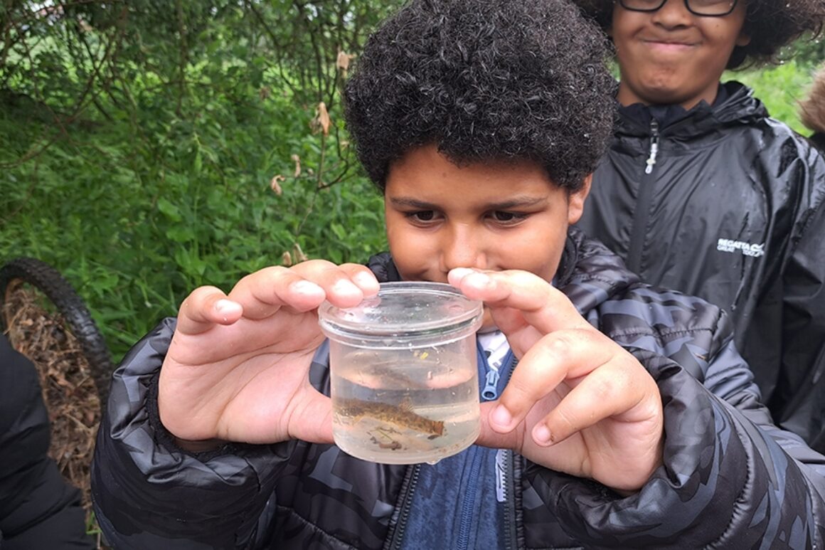 A young person stood on riverbank holding up a plastic magnifying jar containing a small fish, probably stickleback.