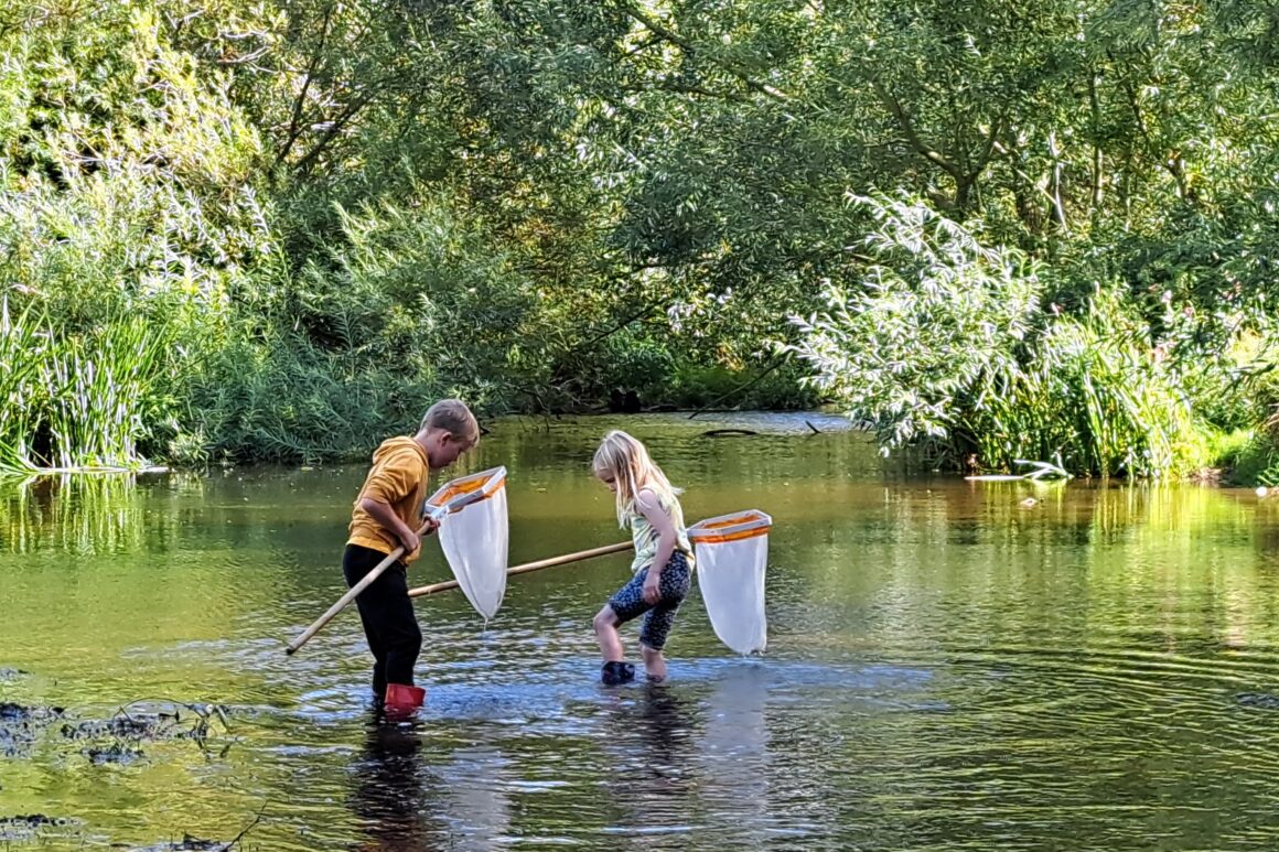 Two young people in wellingtons are stood in a shallow section of river each holding a small 'keep' net. Calm section of river with a backdrop of green trees and foliage in dappled sunlight