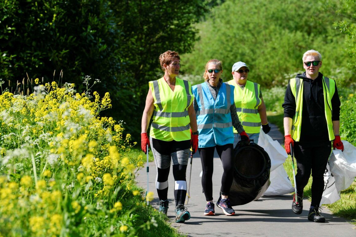A group of four people in high vis vests walking along a tarmac path holding bags full of litter and litter grabbing claws.