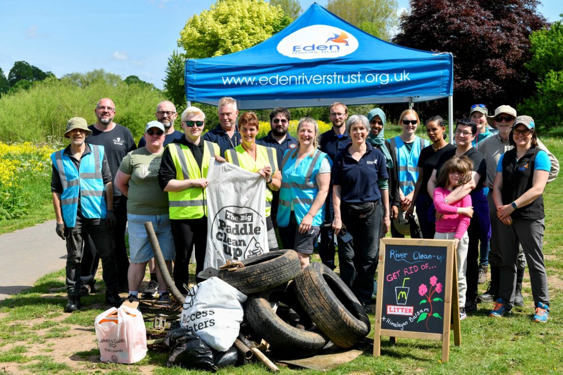 Group of volunteers in blue and yellow hi vis vests stood in front of Eden Rivers Trust gazebo on grass in sunshine. There is a large pile of collected litter in front of them including full back bin bags and a number of car tyres. A small A Board sign reads:
