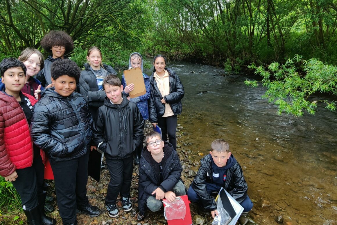 A group of young people stood on the bank of a shallow section of river are posting for a photograph, in coats and looking wet from the rain.