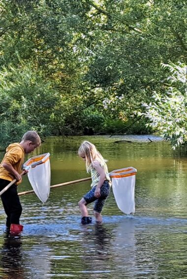 Two young people in wellingtons are stood in a shallow section of river each holding a small 'keep' net. Calm section of river with a backdrop of green trees and foliage in dappled sunlight