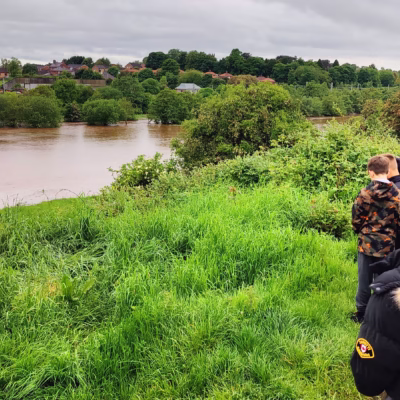 A group of young people stood at the top of a grassy river bank looking towards the river Petteril in high spate following heavy rainfall.