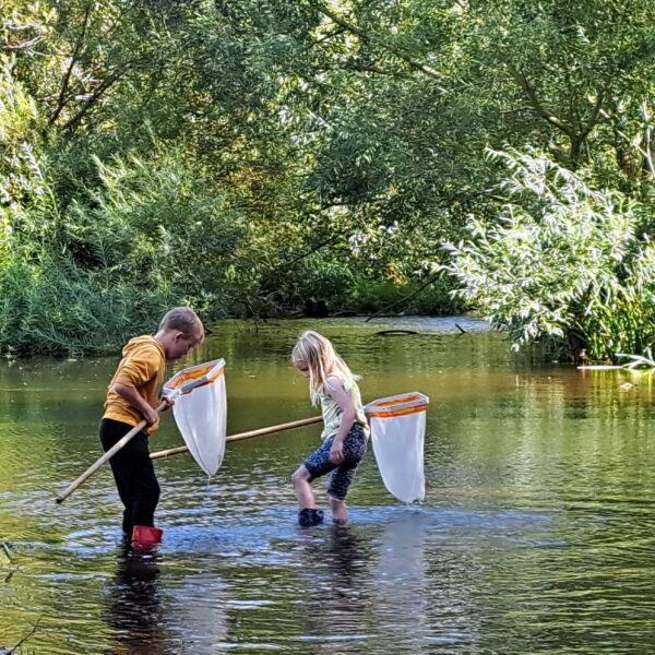 Two young people in wellingtons are stood in a shallow section of river each holding a small 'keep' net. Calm section of river with a backdrop of green trees and foliage in dappled sunlight