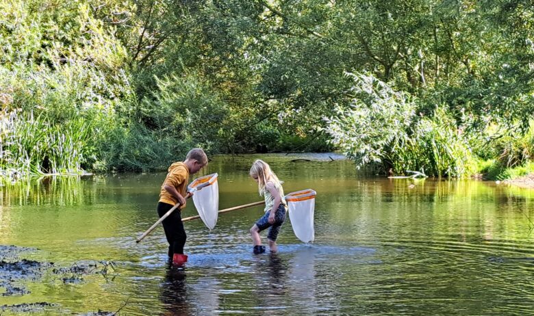 Two young people in wellingtons are stood in a shallow section of river each holding a small 'keep' net. Calm section of river with a backdrop of green trees and foliage in dappled sunlight