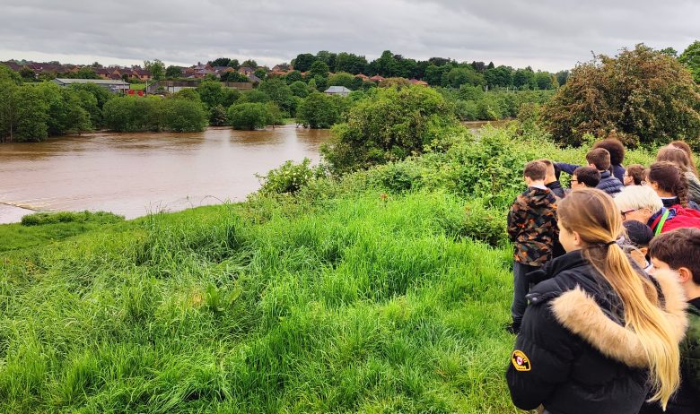 A group of young people stood at the top of a grassy river bank looking towards the river Petteril in high spate following heavy rainfall.