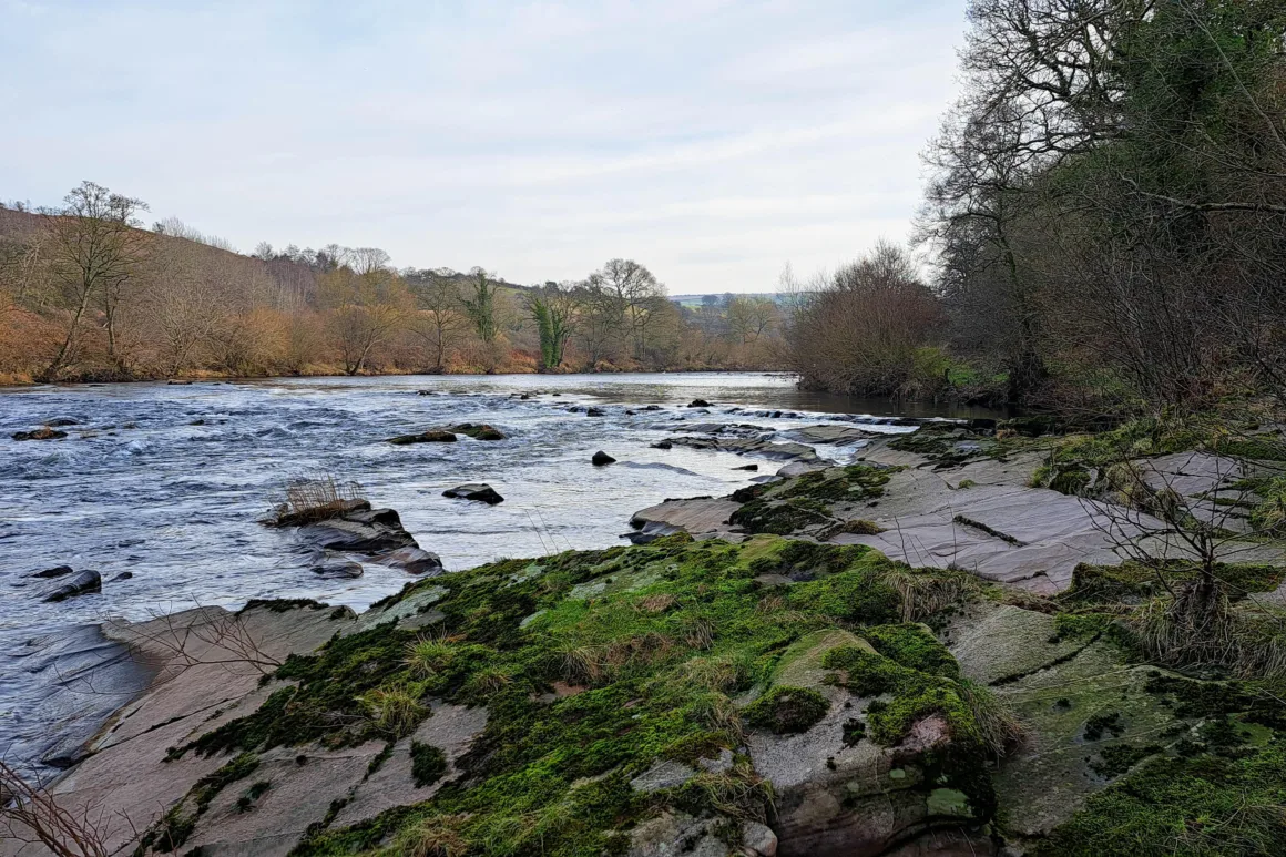 View of a wide section of fast flowing river with large rocky outcrops and limestone boulders to the shore.