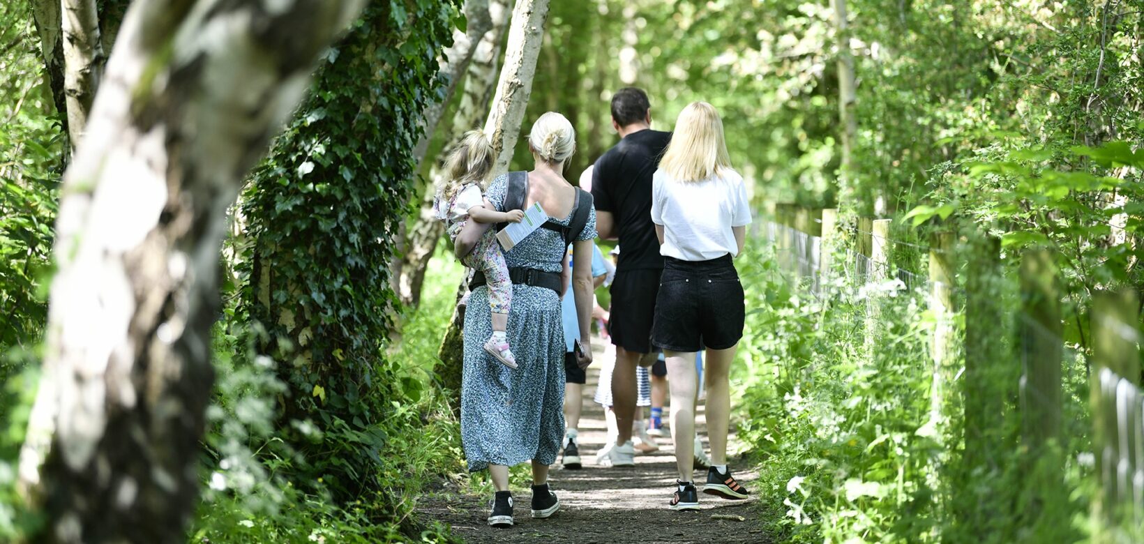 Two men and a woman are walking along a wooded path. One of the women has a toddler balanced on her hip as she walks.
