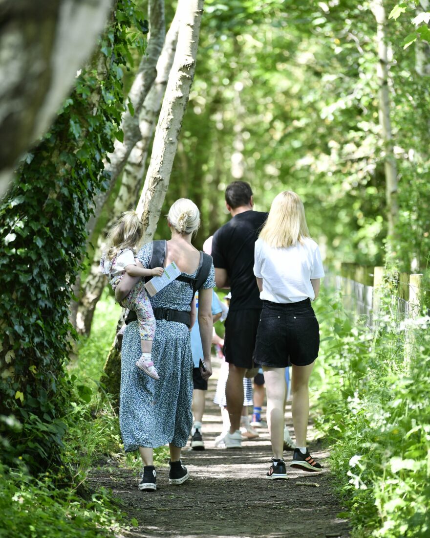 Two men and a woman are walking along a wooded path. One of the women has a toddler balanced on her hip as she walks.