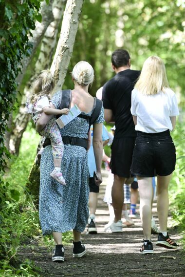 Two men and a woman are walking along a wooded path. One of the women has a toddler balanced on her hip as she walks.