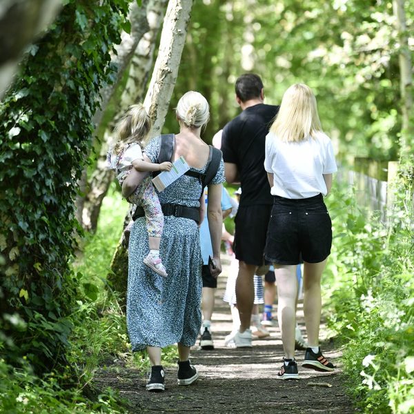 Two men and a woman are walking along a wooded path. One of the women has a toddler balanced on her hip as she walks.