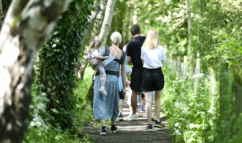 Two men and a woman are walking along a wooded path. One of the women has a toddler balanced on her hip as she walks.