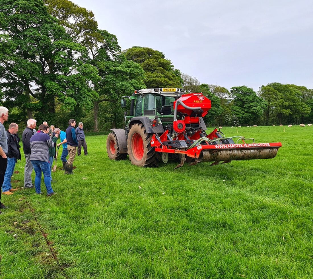 A group of people are gathered in a group on grassland next to a tractor waiting to give them a demonstration of soil aeration.