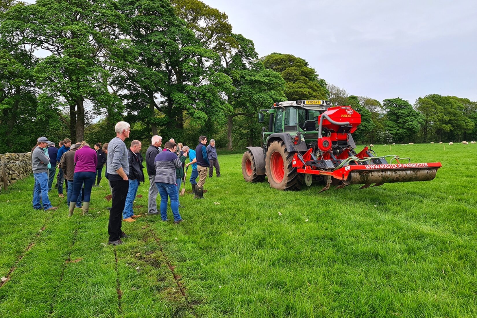 A group of people are gathered in a group on grassland next to a tractor waiting to give them a demonstration of soil aeration.
