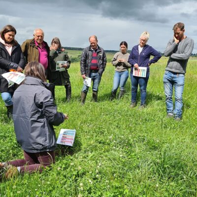 A group of people standing on grassland and arranged in a semi circle to listen to information being given by the activity leader about soil health.