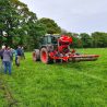 A group of people are gathered in a group on grassland next to a tractor waiting to give them a demonstration of soil aeration.