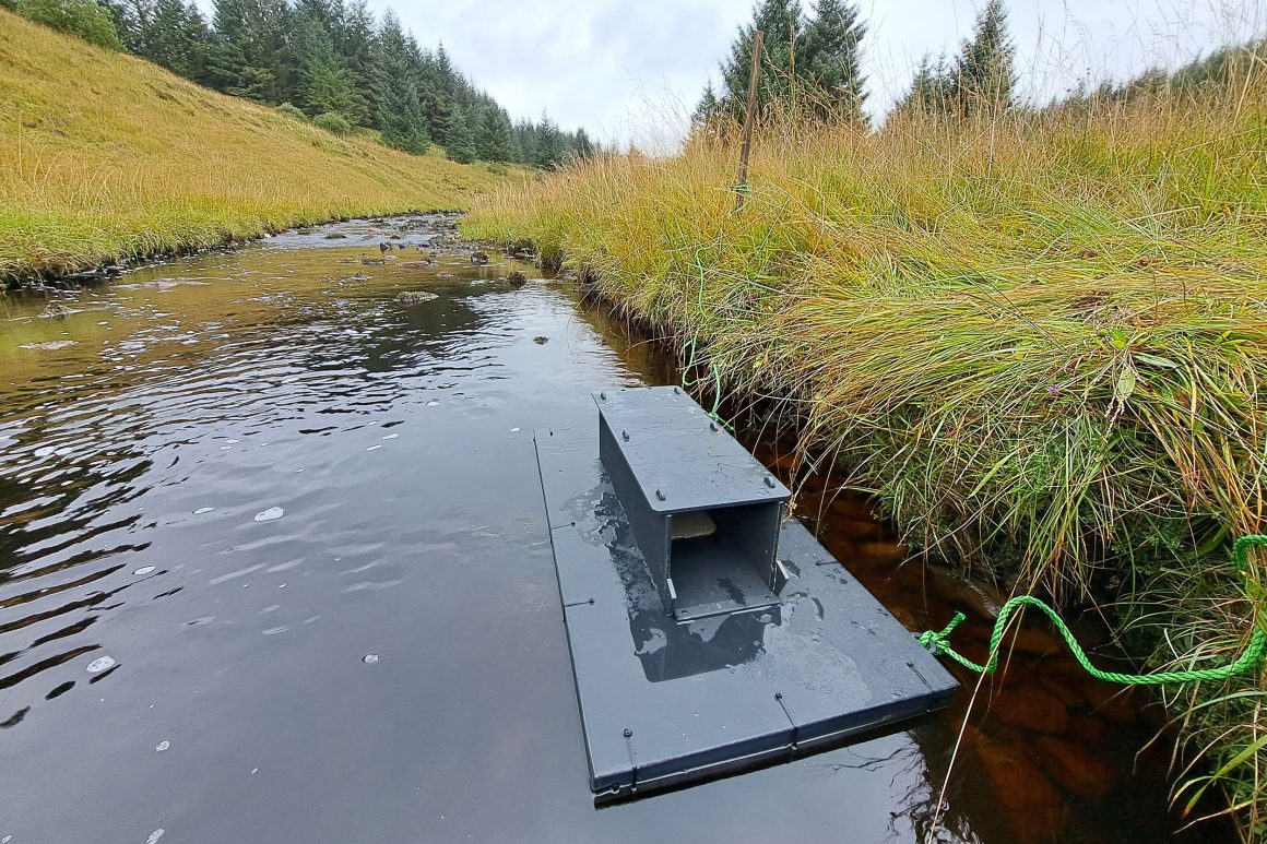 In a valley bottom there is a shallow river that sweep across the landscape. In the foreground there is a Mink raft tethered to the side of the watercourse beneath a grass rich bank.
