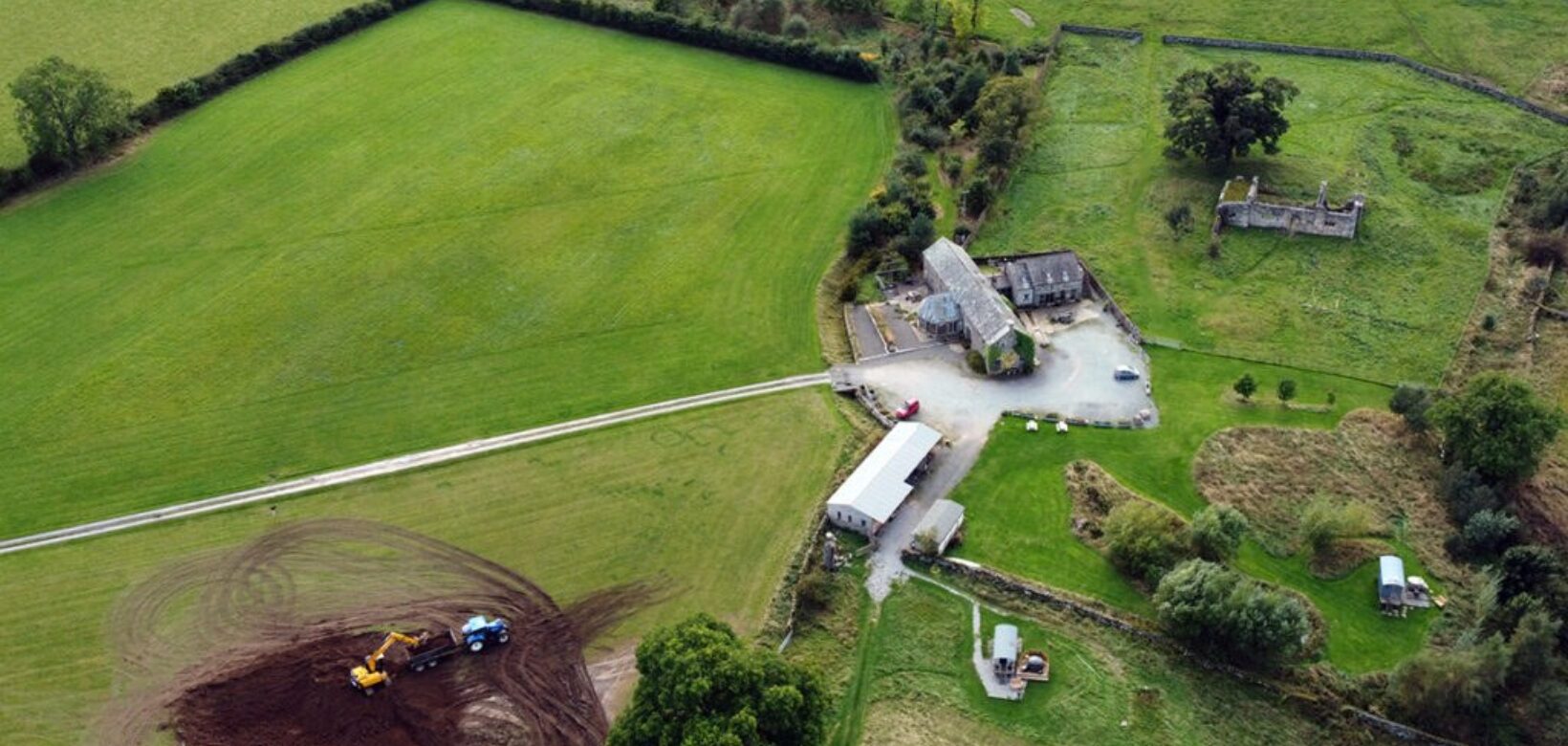 Aerial view of a large circular scrape being excavated by digger on green pastureland.