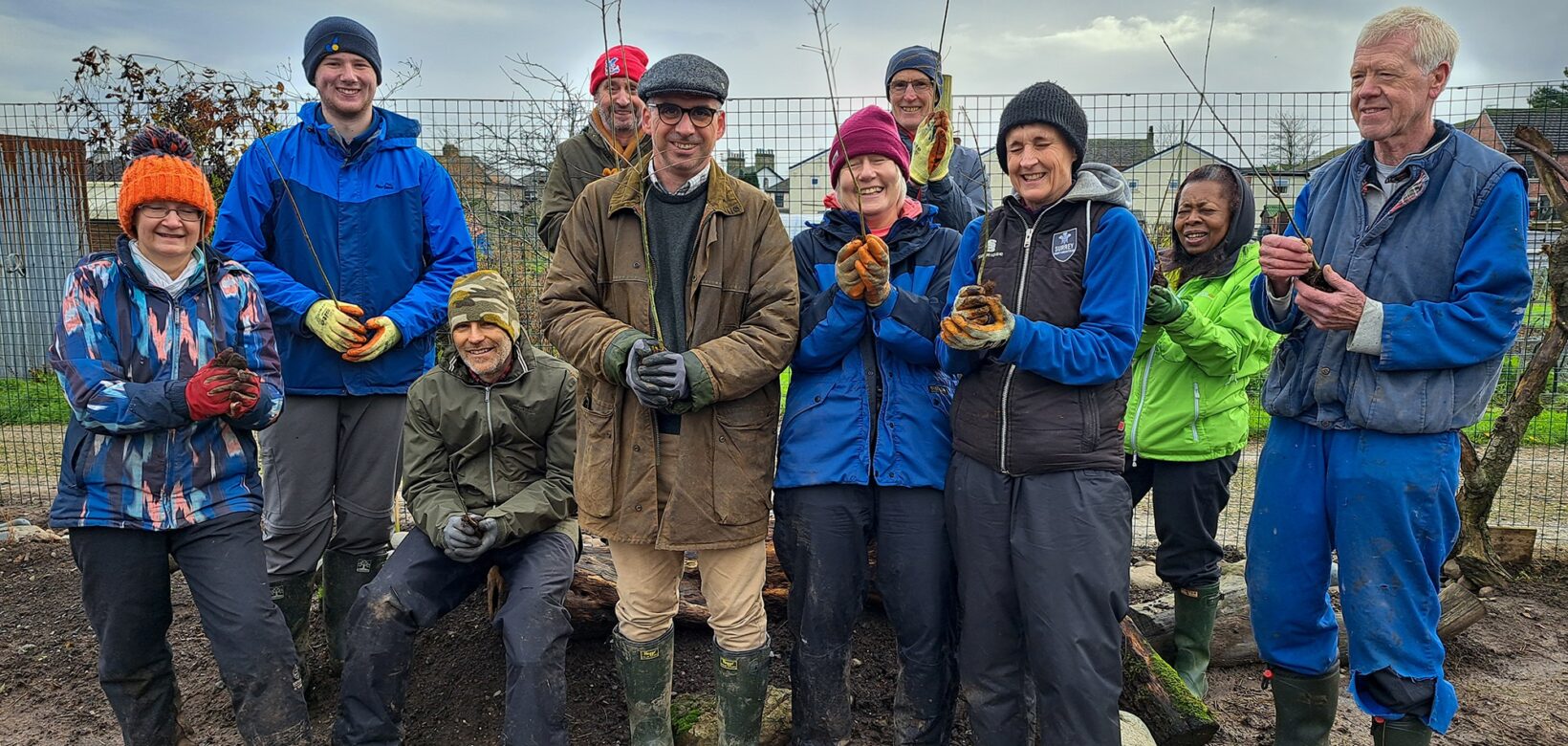 A group of volunteers stand on muddy ground to pose for a photograph, each holding a tree sapling aloft.