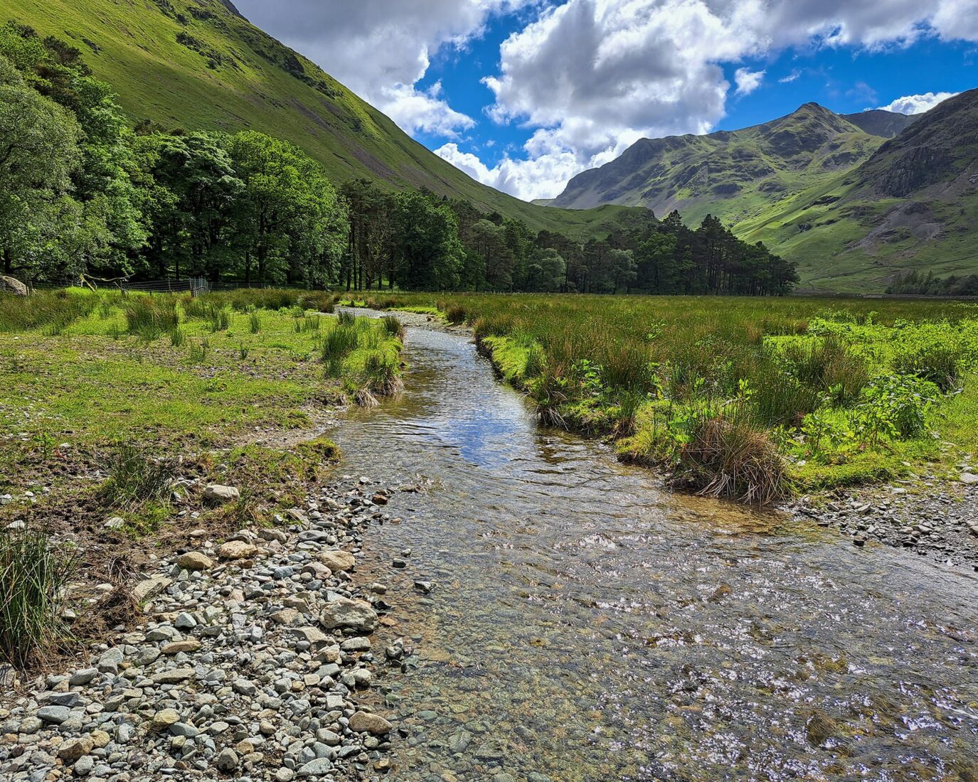 Sunn day. In the background are fells, sweeping away towards the sky. IN the foreground can see clear water running through a pebbly river channel, with wetland grasses scattered on either side of the river on a green field.