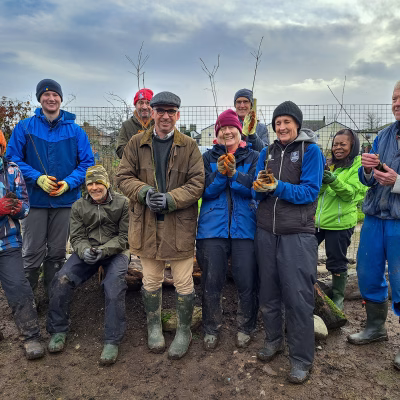 A group of volunteers stand on muddy ground to pose for a photograph, each holding a tree sapling aloft.