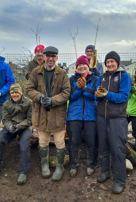 A group of volunteers stand on muddy ground to pose for a photograph, each holding a tree sapling aloft.